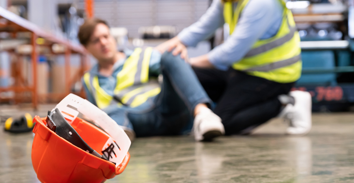 Ein Arbeiter ist hingefallen. Ein anderer kommt diesem zur Hilfe. Im Vordergrund ist ein Schutzhelm zu sehen.