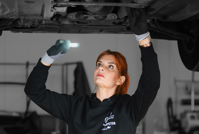 Die Kfz-Mechatronikerin Anne Althaus-Braun inspiziert den Unterbodens eines aufgebockten Autos mit einer Leuchte.