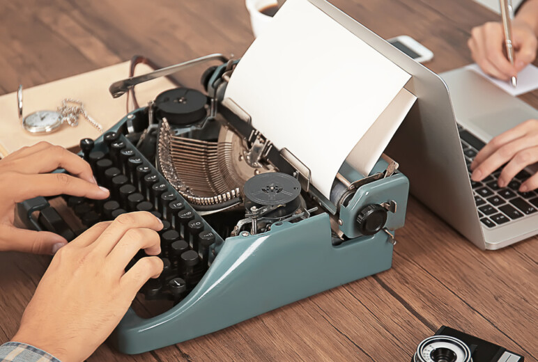 Detailaufnahme von Arbeitsplatz: Tisch mit Schreibmaschine und Laptop gegenüber mit tippenden Händen