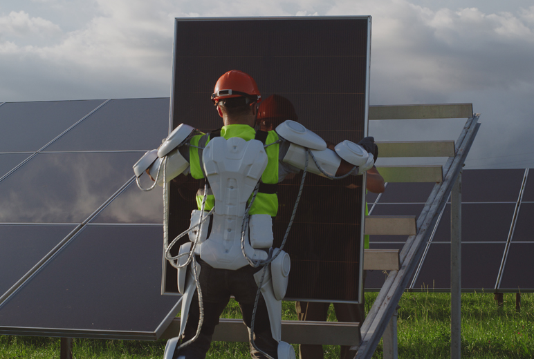 Person mit Exoskelett auf einer Wiese, die eine Solarpanele hält, dahinter ein schräges Gestell zur Anbringung. Daneben und davor stehen weitere schon montierte, in etwa 90 Grad aufgerichtete Solarpanelen auf Gras.