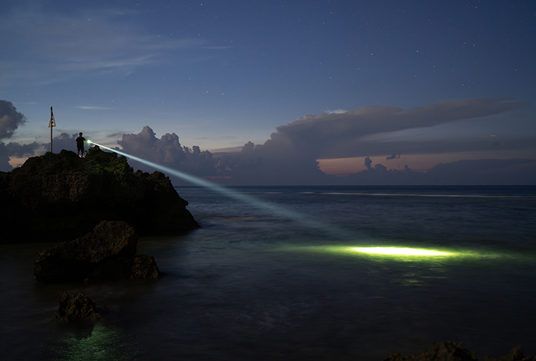 Eine Person steht bei Dämmerung oder Nacht auf einem Felsen am Meer und leuchtet mit einer leistungsstarken Taschenlampe über das Wasser. Der Lichtstrahl ist deutlich sichtbar und trifft weit draußen auf die Wasseroberfläche. Am Himmel sind Sterne und Wolken zu erkennen.