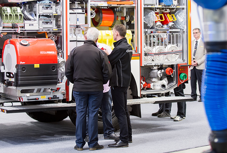 Mehrere Personen stehen vor einem geöffneten Einsatzfahrzeug der Feuerwehr, das mit verschiedenen technischen Geräten und Ausrüstungsgegenständen ausgestattet ist. Ein Fachmann erklärt den Anwesenden Details der Ausrüstung. Im Hintergrund beobachtet ein Mann im Anzug die Szene. Die Umgebung deutet auf eine Messe oder Fachausstellung hin.