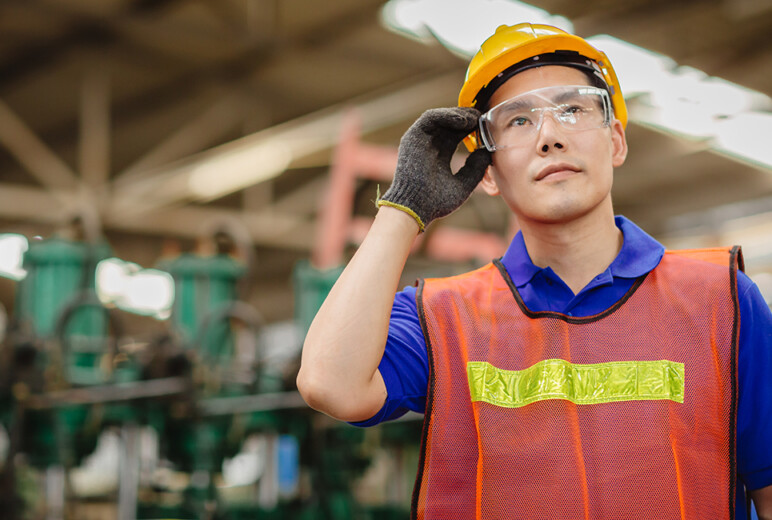 Ein Mann mit Schutzhelm, Schutzbrille und Warnweste steht in einer Industriehalle und fässt sich an die Brille.