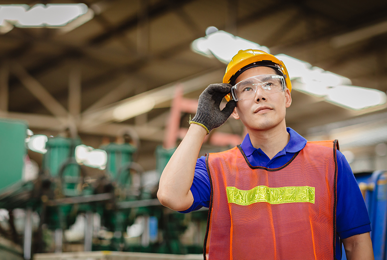 Ein Mann mit Schutzhelm, Schutzbrille und Warnweste steht in einer Industriehalle und fässt sich an die Brille.