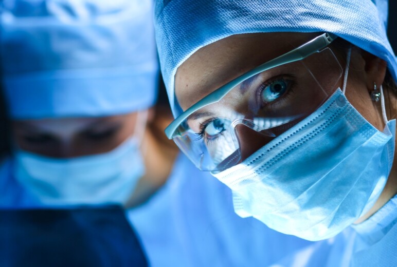 Zwei Personen in OP-Kleidung und mit Mundschutz, von denen die Frau im Vordergrund eine Schutzbrille trägt und direkt in die Kamera blickt.