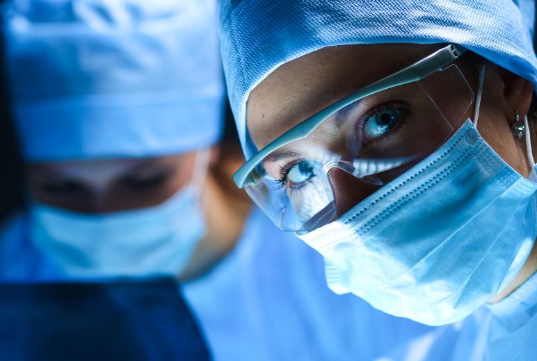 Zwei Chirurginnen in OP-Kleidung und Mundschutz arbeiten konzentriert in einem Operationssaal. Die Frau im Vordergrund trägt zusätzlich eine Schutzbrille und blickt direkt in die Kamera.