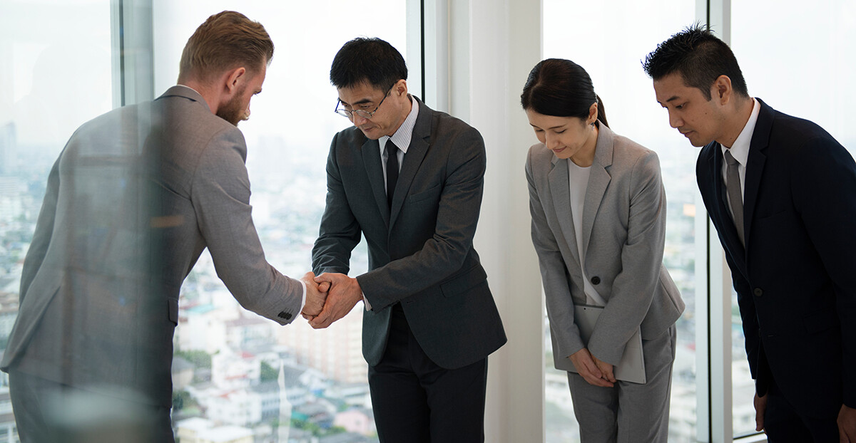 Vier Geschäftsleute in formeller Kleidung begrüßen sich in einem modernen Büro mit großen Fenstern. Ein westlicher Mann und ein asiatischer Mann geben sich die Hand, während sie sich gegenseitig respektvoll verbeugen. Zwei weitere asiatische Kollegen, eine Frau und ein Mann, stehen daneben und verbeugen sich ebenfalls leicht.