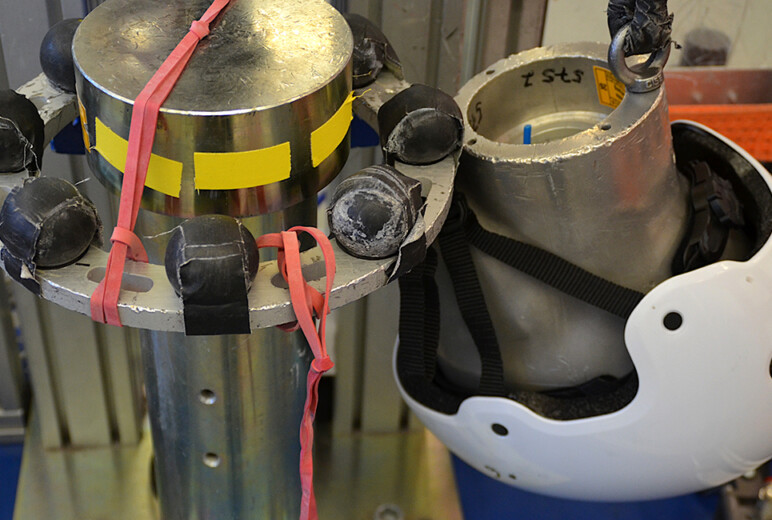 Ein Prüfstands zur Helmsicherheit. Links ist ein zylindrischer Metallpfosten mit schwarzen Sensoren und roten Gummibändern zu sehen. Rechts daneben hängt ein weißer Schutzhelm an einem Aluminiumkopfmodell, das an einem Seil befestigt ist. Im Hintergrund sind Metallrahmen und orangefarbene Sicherungsbänder sichtbar.