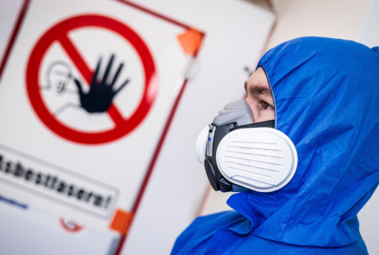 Eine Person im blauen Einwegoverall trägt eine Halbmaske mit Partikelfiltern und steht vor einer Tür mit durchgestrichenem Handsymbol und dem Warnhinweis „Asbestfasern!“.