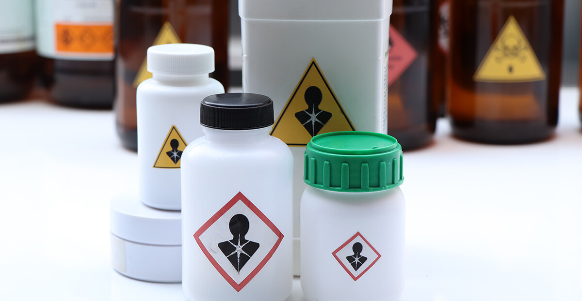 Mehrere weiße Kunststoffbehälter und braune Glasflaschen stehen dicht beieinander auf einer weißen Fläche, alle mit dem GHS-Piktogramm „Gesundheitsgefahr“ gekennzeichnet.