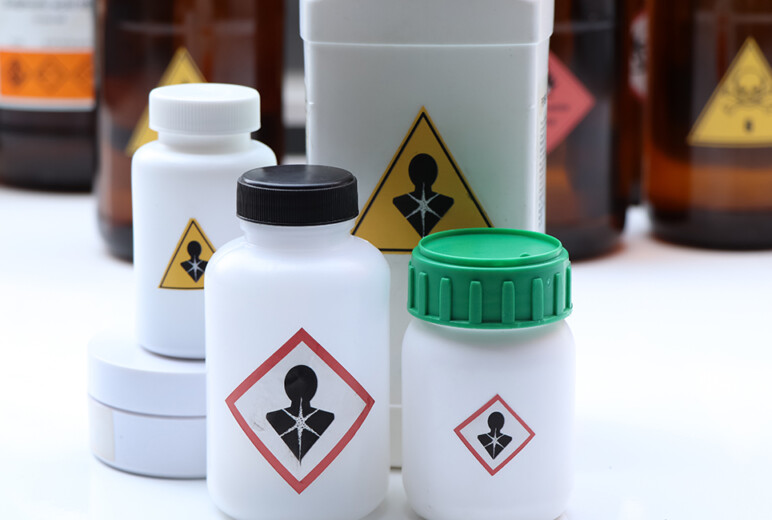 Mehrere weiße Kunststoffbehälter und braune Glasflaschen stehen dicht beieinander auf einer weißen Fläche, alle mit dem GHS-Piktogramm „Gesundheitsgefahr“ gekennzeichnet.