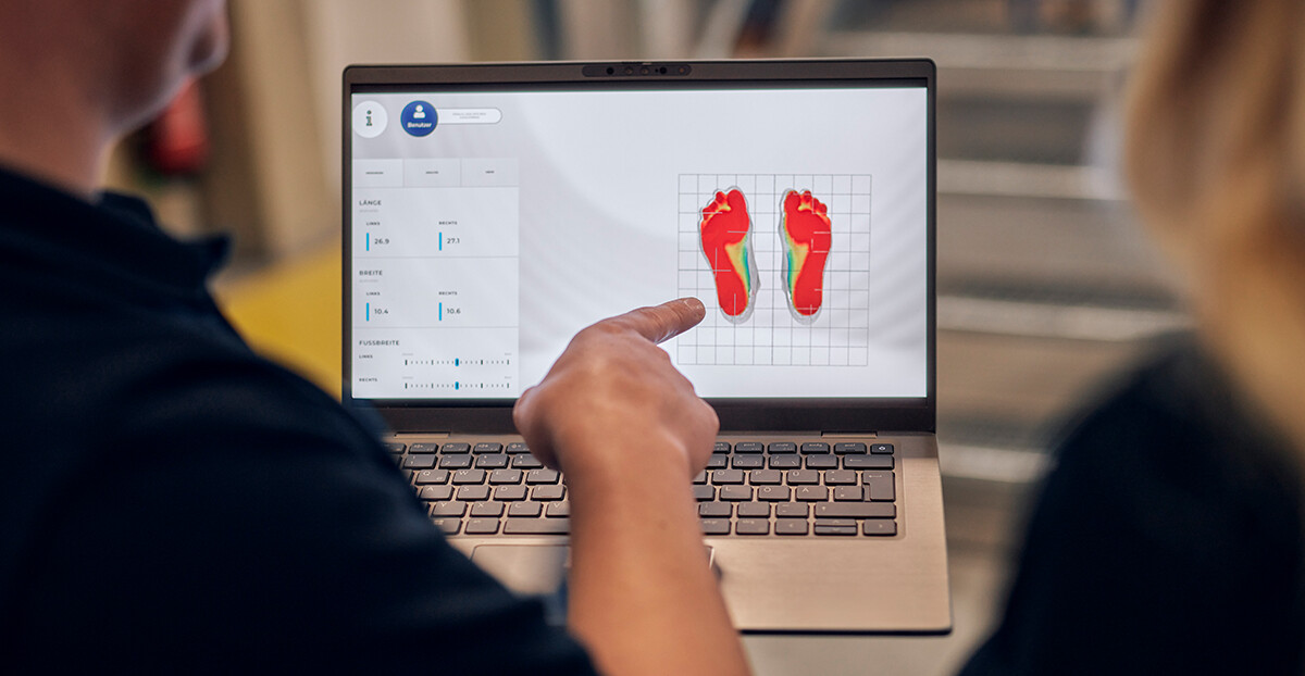 Ein Laptopbildschirm zeigt farbige Druckverteilungen beider Füße, eine Person deutet mit dem Finger darauf, während eine zweite Person zusieht.