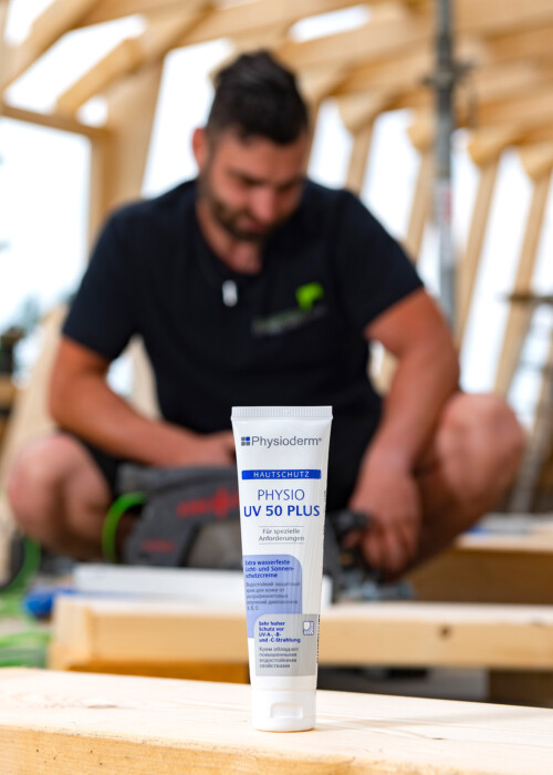 Eine Tube Physioderm UV 50 Plus steht im Vordergrund einer Holzbaustelle, im unscharfen Hintergrund arbeitet ein Handwerker.