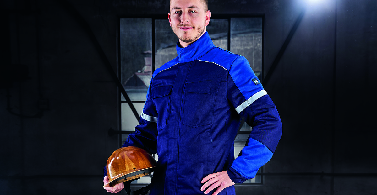 Ein Mann in dunkelblauer Schutzkleidung mit Reflexstreifen hält einen gelben Schutzhelm in der Hand und steht in einer schwach beleuchteten Industriehalle.