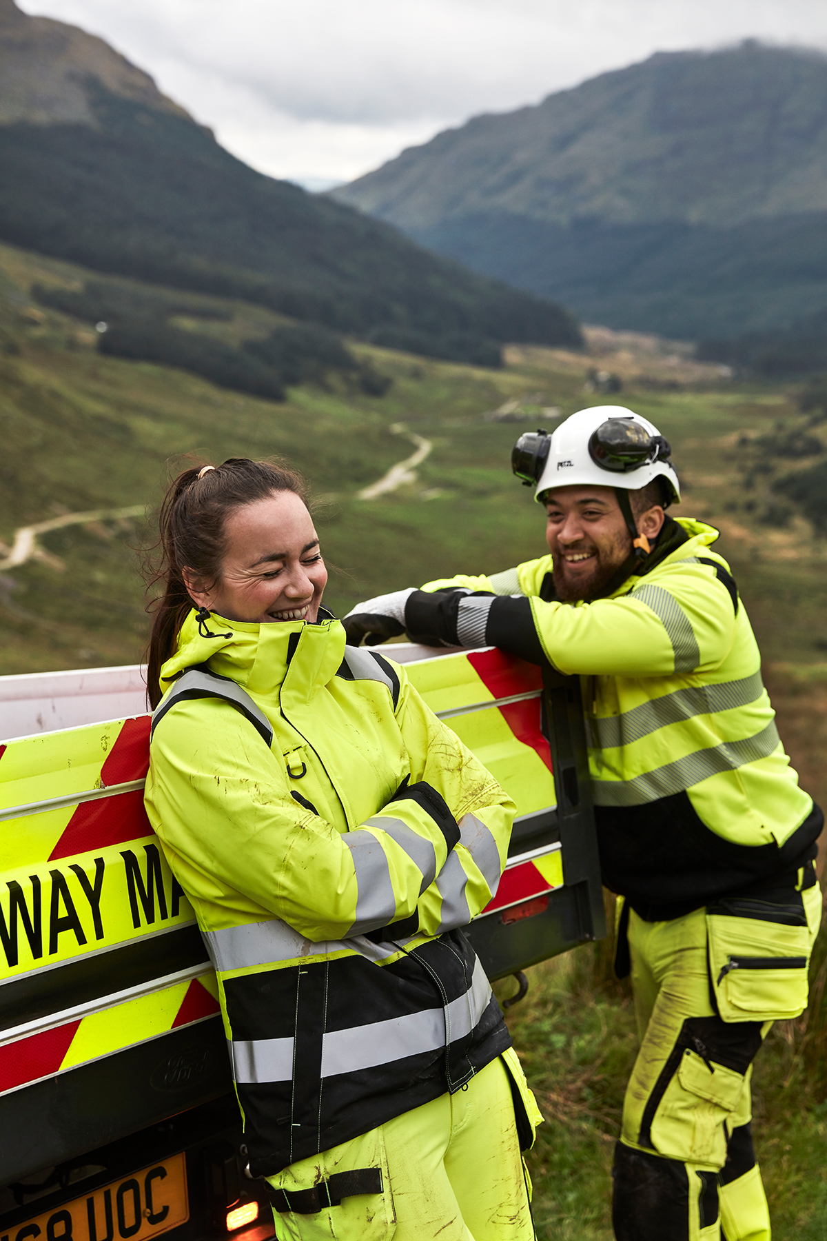 Eine Arbeiterin und ein Arbeiter in gelben Warnjacken lehnen lachend an ein Arbeitsauto vor grüner Landschaft. Der Mann trägt einen weißen Schutzhelm.