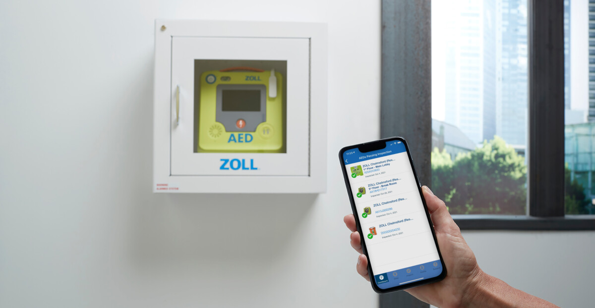 Ein automatischer externer Defibrillator in einem weißen Wandschrank der Marke Zoll ist durch das Sichtfenster sichtbar, davor hält eine Hand ein Smartphone mit der dazugehörigen App.