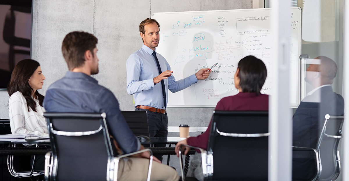 Ein Mann präsentiert vor einem Whiteboard mit Diagrammen und handschriftlichen Notizen, während vier Kolleginnen und Kollegen zuhören.