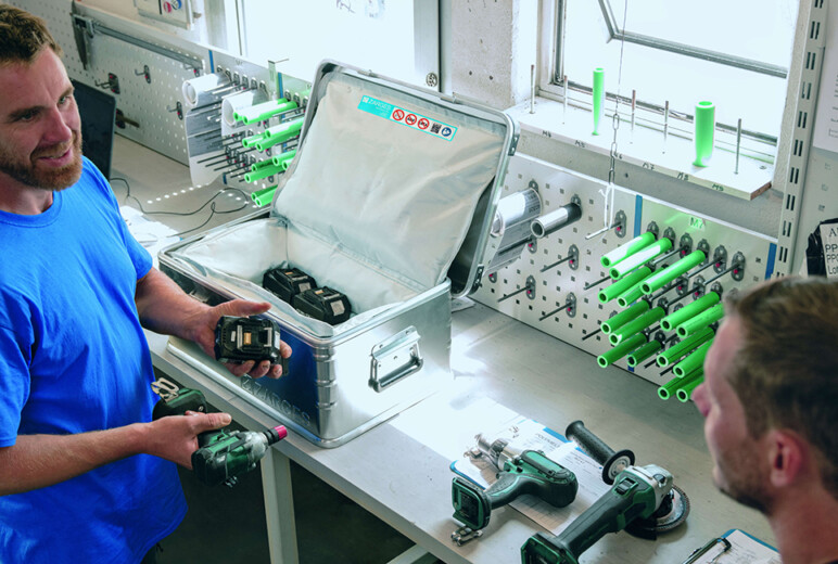 In einer Werkstatt hält ein Mitarbeiter im blauen Shirt einen Akkupack hoch, auf der Werkbank stehen ein offener Aluminiumkoffer mit weiteren Lithium-Batterien und mehrere Elektrowerkzeuge.