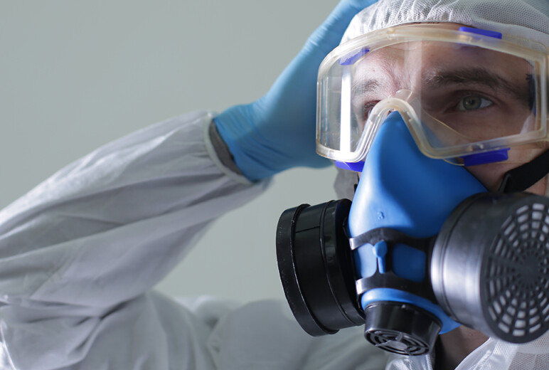 Eine Person in Schutzanzug und blauen Einweghandschuhen trägt eine Vollgesichtsatemschutzmaske mit Filtern und hält beide Hände an den Kopf.