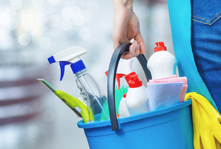 Person trägt einen blauen Putzeimer mit Sprühflasche, Reinigungsmitteln, Tuch und gelben Gummihandschuhen.