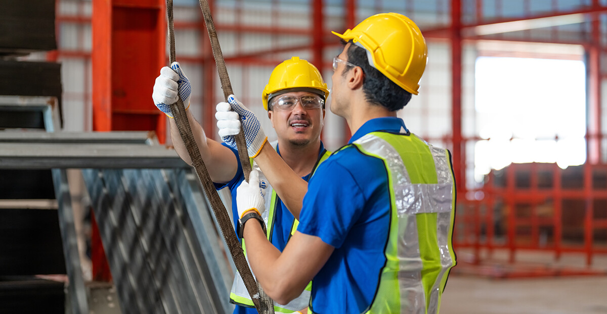Zwei Bauarbeiter tragen Sicherheitshelme und Schutzwesten. Beide halten ein Seil und unterhalten sich. Die Szene findet auf einer Baustelle statt.