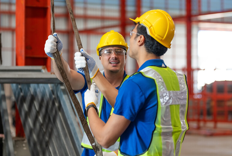 Zwei Bauarbeiter tragen Sicherheitshelme und Schutzwesten. Beide halten ein Seil und unterhalten sich. Die Szene findet auf einer Baustelle statt.