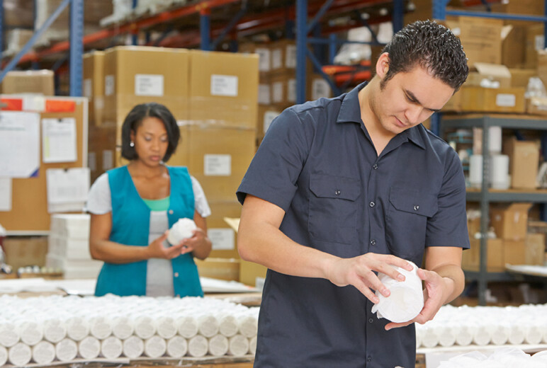 Zwei Beschäftigte prüfen verpackte Produkte an einem Arbeitstisch im Warenlager; im Hintergrund stehen hohe Regale und gestapelte Kartons.