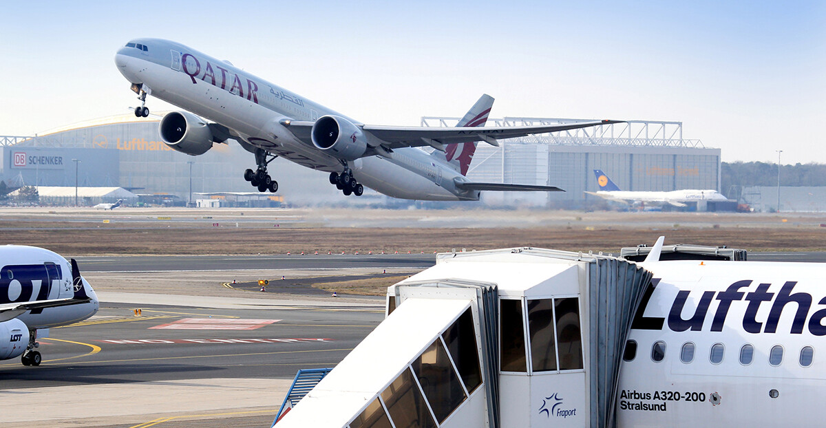 Am Flughafen hebt ein Qatar-Airways-Jet ab, im Vordergrund steht ein Lufthansa-Flugzeug mit angedockter Fluggastbrücke.