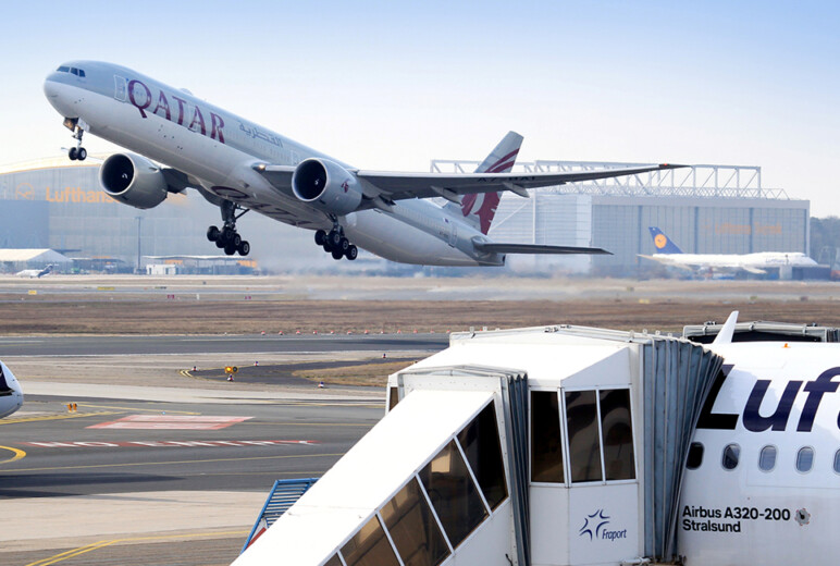 Am Flughafen hebt ein Qatar-Airways-Jet ab, im Vordergrund steht ein Lufthansa-Flugzeug mit angedockter Fluggastbrücke.