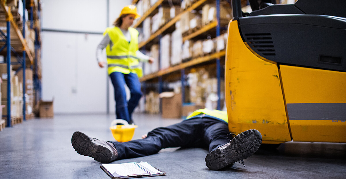 In einer Lagerhalle liegt ein Arbeiter vor einem Gabelstapler am Boden, eine Kollegin in Warnweste eilt herbei, der gelbe Helm liegt daneben.